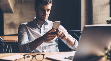 Homem sentado em um escritório, usando um celular em frente ao notebook