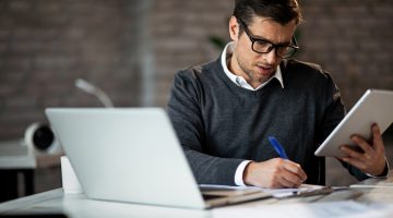 homem em frente a um notebook e escrevendo em um caderno enquanto realiza seu quiz tributário