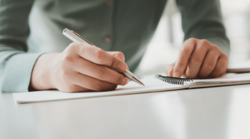 mãos segurando uma caneta e escrevendo em um caderno, fazendo menção ao planejamento anual